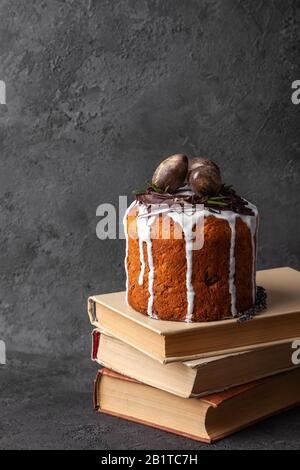 Kulich baked Easter cake with white glaze decorated sweet eggs in russian orthodox tradition stands on old books, vertical image, dark background, cop Stock Photo