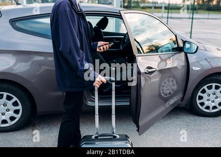 Unrecognizable man standing at car with suitcase and holding cell phone Stock Photo