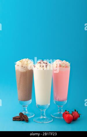 Three milkshakes - vanilla strawberry chocolate in high transparent glasses, whipped cream, cookies, on blue background, vertical Stock Photo