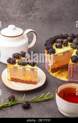 Piece of fresh blackberry cake on platter, covered with purple cream, caramel, flows down on the sides, teapot and tea cup on gray surface, vertical Stock Photo