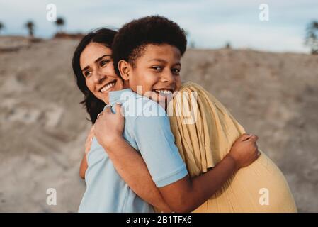 Close up of young mother and happy son embracing on beach at sunset