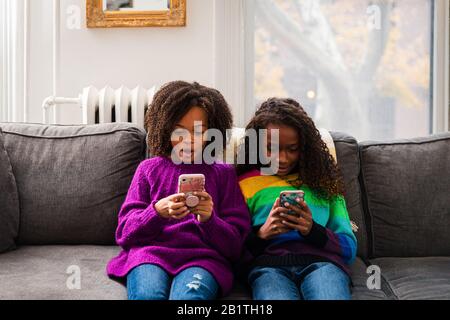 Sisters in sweaters using mobile phones while sitting on sofa at home Stock Photo