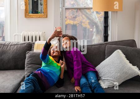 Smiling girl taking selfie with sister while leaning on sofa at home Stock Photo
