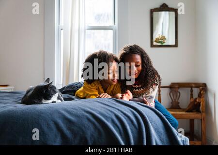 Smiling sisters using smart phone while lying by cat on bed in bedroom Stock Photo