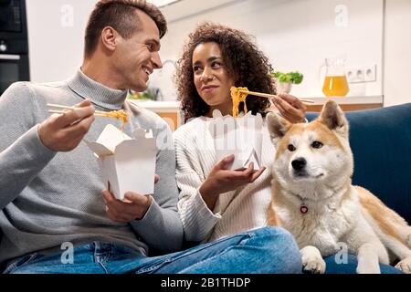 Romantic Date. Young multiethnic couple at home sitting on sofa with dog eating noodle talking smiling happy Stock Photo