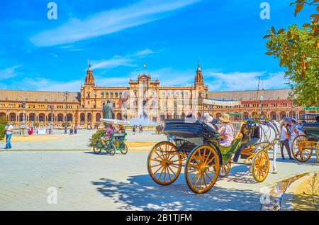 SEVILLE, SPAIN - OCTOBER 1, 2019: The line of tourist horse-drawn carriages on the shade of trees of Maria Luisa park with a view on Plaza de Espana, Stock Photo