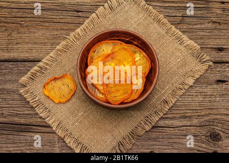 Dried slices of persimmon in bowl. Fruit snack, healthy eating concept. Sackcloth, old wooden boards background, top view, copy space Stock Photo