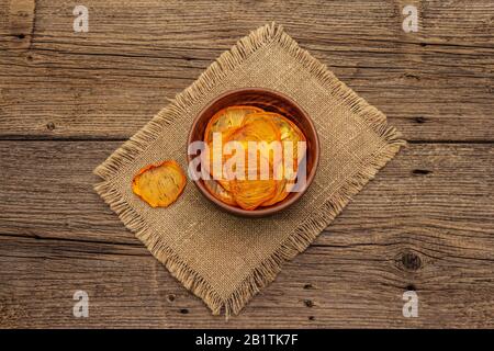 Dried slices of persimmon in bowl. Fruit snack, healthy eating concept. Sackcloth, old wooden boards background, top view, copy space Stock Photo