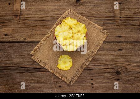 Dried slices of pineapple in bowl. Fruit snack, healthy eating concept. Sackcloth, old wooden boards background, top view, copy space Stock Photo