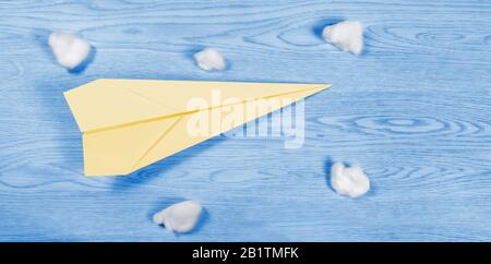 Yellow paper airplane among cotton wool balls over a wooden blue background resembling a blue sky and a few clouds Stock Photo