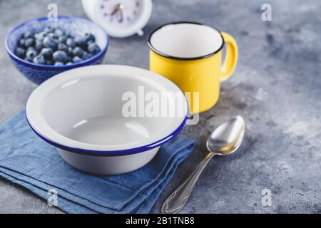Empty metal bowl for breakfast, enamel yellow cup, blueberry, alarm clock and spoon on blue napkin, copy space. Stock Photo