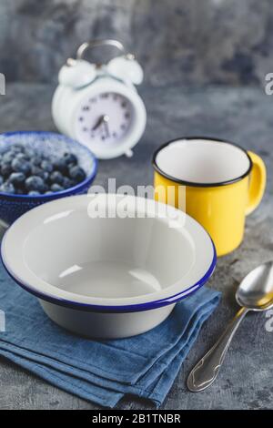 Empty metal bowl for breakfast, enamel yellow cup, blueberry, alarm clock and spoon on blue napkin, copy space. Stock Photo