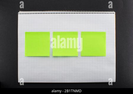 Blank notepad with three empty green sticky notes on black background. Empty notebook with white texture and brown grid lines. Copy space for add text Stock Photo