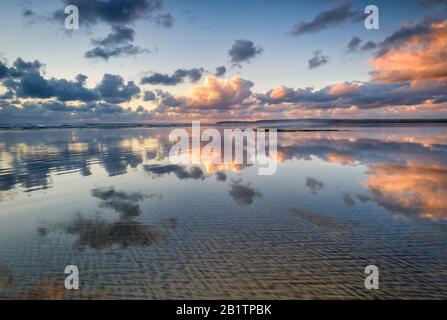 Mirror image of the sun setting and the clouds reflecting on the wet sand of the beach, coastal living, dramatic skies, reflections, sand, atmospheric Stock Photo