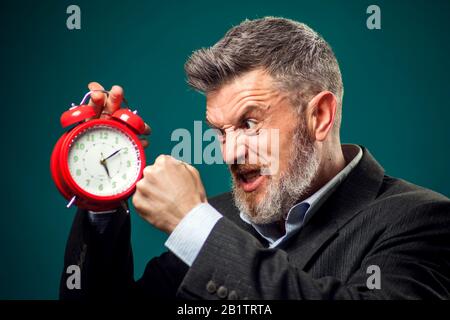 Angry bearded businessman in suit holding red alarm clock and shouting at it. Time management concept Stock Photo
