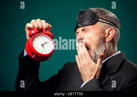 Bearded businessman in suit witn sleep mask on head holding red alarm clock and yawns, wants sleep. Time management concept Stock Photo