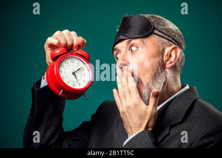 Bearded businessman in suit witn sleep mask on head holding red alarm clock and yawns, wants sleep. Time management concept Stock Photo