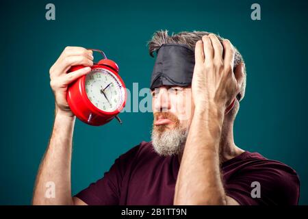 Surprised man in red t-shirt and sleep mask on head holding red alarm clock. Lifestyle and bed time concept Stock Photo