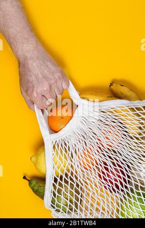 Zero waste plastic free concept. A human hand holds a string bag with fresh fruits on yellow paper background. Mesh shopping bag with fruits. Sustaina Stock Photo