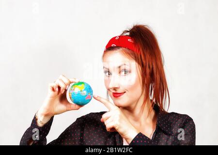 Young caucasian happy redhead pin up girl with red lips and bandana showing little earth symbol and looking for holiday destination Stock Photo