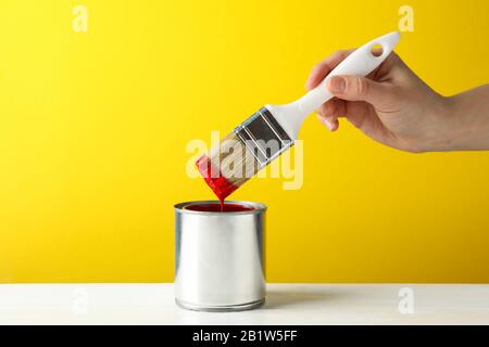 Female hand holding brush over the can with red paint against yellow background Stock Photo