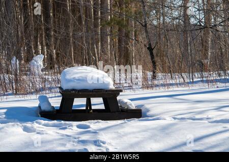 A wooden picnic table is covered in snow in the winter. The area has not been cleared after a snowfall, and a thick layer of snow remains on the table Stock Photo