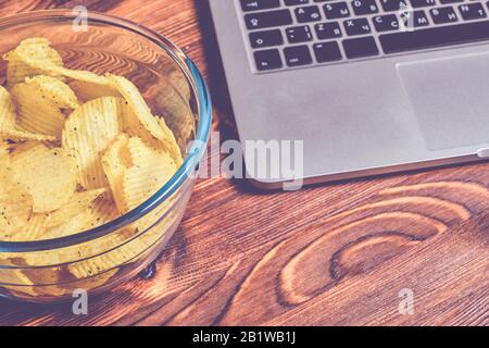 Fluted chips in a glass bowl next to a laptop on a wooden table. The concept of rest. Toned. Close up. Stock Photo