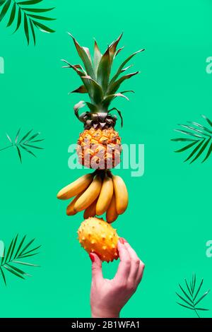 Assortment of tropical fruits, pyramid balancing o human hand on green background. Pineapple, kiwano, kiwi , lichee, banana - tower made of exotic fru