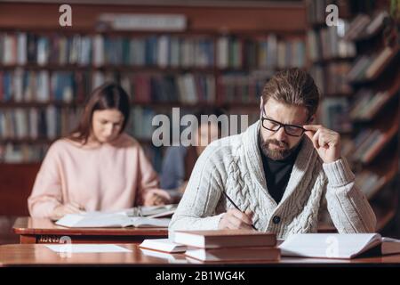 Mature bearded man in casual clothing and eyeglasses sitting at table with lots of book at public library. Serious male searching for information. Edu Stock Photo