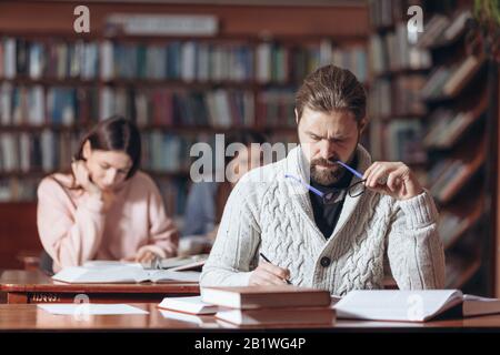 Concentrated unshaven man in knitted sweater and eyeglasses taking notes while reading books at public library. Mature male sitting at table with blur Stock Photo