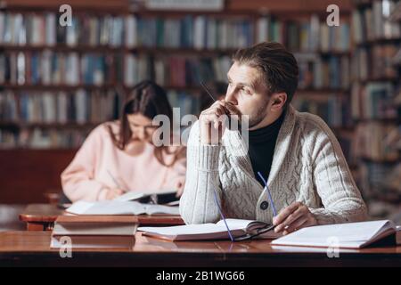 Bearded male reader in casual clothing and eyeglasses sitting at table at public library and looking away. Mature man reading books and taking notes. Stock Photo