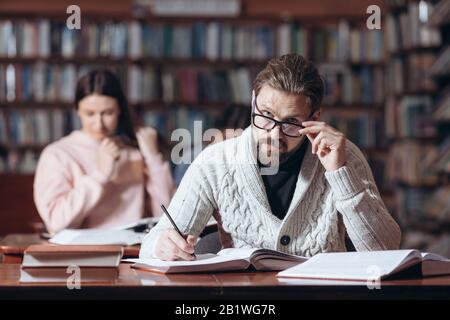 Confident middle-aged man in knitted sweater and eyeglasses taking notes while reading books at library. Bearded reader looking at camera while sittin Stock Photo