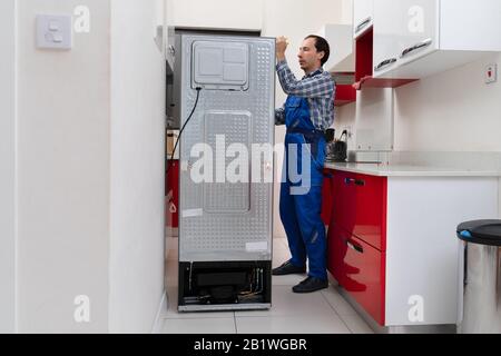 Young Male Serviceman Repairing Broken Refrigerator In Kitchen Stock Photo