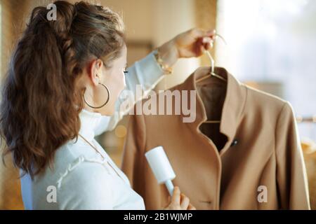 Preparing for cold season. Seen from behind trendy 40 years old housewife in white sweater and skirt cleaning coat on hanger with lint roller in the m Stock Photo