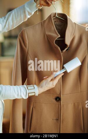 Preparing for cold season. Closeup on woman in white sweater cleaning coat on hanger with lint roller in the house in sunny winter day. Stock Photo