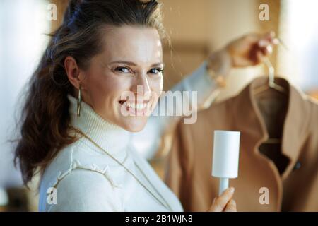 Preparing for cold season. Portrait of happy middle age housewife in white sweater and skirt cleaning coat on hanger with lint roller in the modern ho Stock Photo