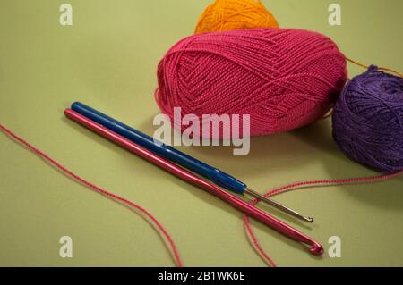 three skeins of yarn of different colors and two hooks of pink and blue on a green background Stock Photo