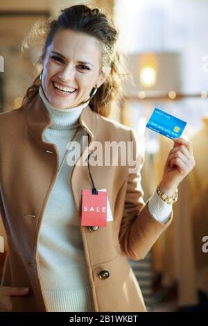 happy elegant woman in white sweater and skirt trying beige coat with red sale price tag and holding blue credit card in modern fashion showroom. Stock Photo