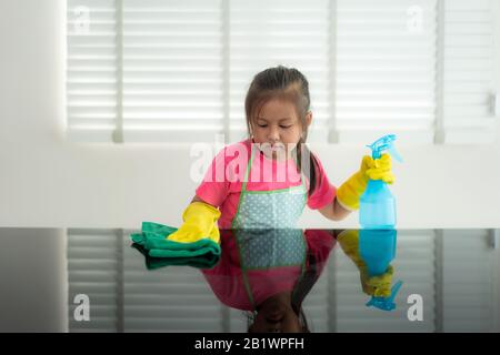 Asian Cute preschool student girl washing glass table with green towel in living room at home, with water and detergent solution spray. Stock Photo