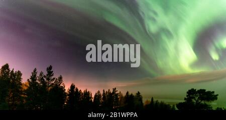 View of brilliant green Aurora shining over Swedish foggy forest landscape in mountains, light rays from a village and Northern Lights color sky in di Stock Photo