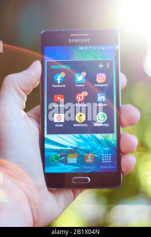 FEBRUARY 3, 2020 - BUENOS AIRES, ARGENTINA: Vertical shot of hand holding a mobile phone showing group of marketing apps in virtual folder with blurre Stock Photo