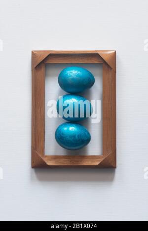 Three blue colored easter eggs placed in the wooden frame on white background. Top view. Easter festive holiday background. Minimalistic photo Stock Photo