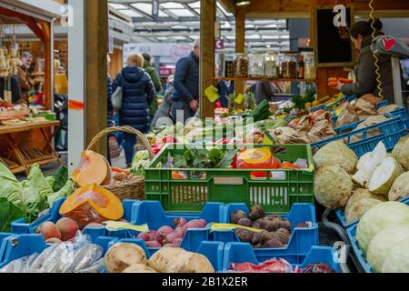 Various vegetables sell on row of blue and green plastic baskets on counter of stall at open air market with blur background of people. Stock Photo