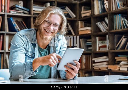 Young bearded man glasses library desk hold modern tablet laugh at camera. Stock Photo