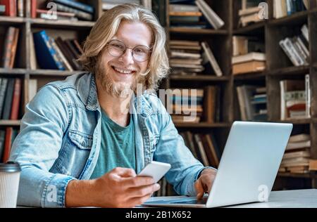 Portrait young man library desk hold smartphone use app laptop education concept. Stock Photo