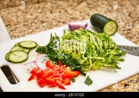 Chopped organic red pepper, parsley, onion, cucumber and romaine lettuce on a white cutting board with cutting knife and shallow depth of field Stock Photo
