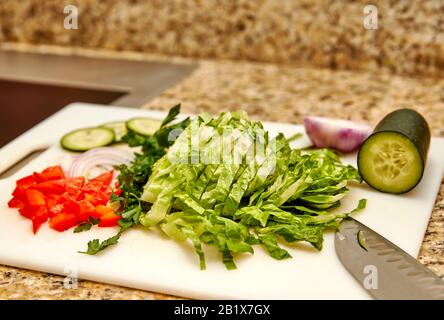 Chopped organic red pepper, parsley, onion, cucumber and romaine lettuce on a white cutting board with cutting knife and shallow depth of field Stock Photo
