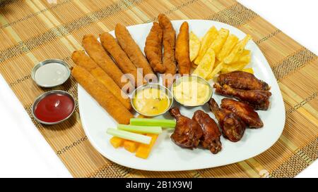 White plate with BBQ chicken wings and breaded chicken fingers accompanied by french fries, carrot and celery pieces, with sauces in small plates on w Stock Photo