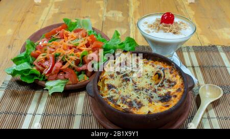 Clay dish with meat lasagna accompanied by carrot, tomato and lettuce salad, and a glass of coconut juice on a wooden table Stock Photo