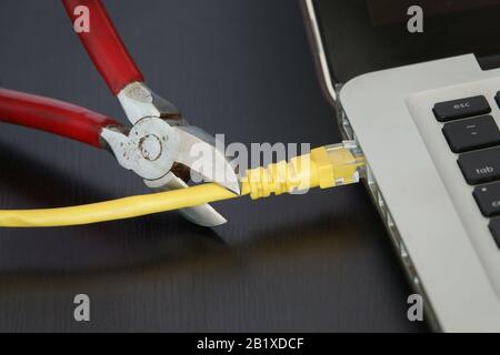 A pair of wire snippers are shown just before cutting a yellow ethernet internet cable connected to a laptop computer. Stock Photo